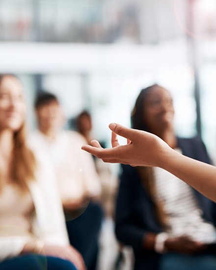 Nahaufnahme einer gestikulierenden Hand einer vortragenden Person; im Hintergrund unscharf ein sitzendes Publikum in einem hellen Seminarraum.