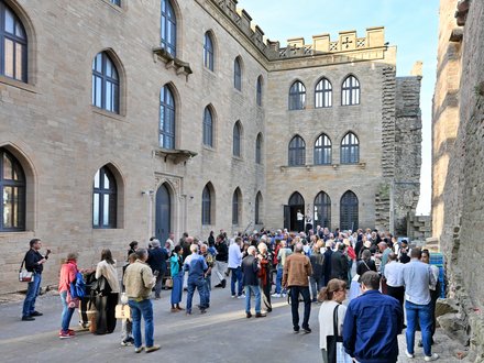 "Große Außenveranstaltung in einem historischen Innenhof mit Backsteingebäuden. Etwa 100 Personen in casual bis Business-Kleidung stehen und gehen durcheinander. Die umgebenden Gebäude zeigen mittelalterliche Architektur mit großen gotischen Fenstern, Zinnen und Steinmauern in Beige und Grau. Sonnenschein bei klarem Himmel beleuchtet die Szene. Links sitzt eine Person mit Tablet und nimmt Notizen."