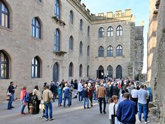 "Große Außenveranstaltung in einem historischen Innenhof mit Backsteingebäuden. Etwa 100 Personen in casual bis Business-Kleidung stehen und gehen durcheinander. Die umgebenden Gebäude zeigen mittelalterliche Architektur mit großen gotischen Fenstern, Zinnen und Steinmauern in Beige und Grau. Sonnenschein bei klarem Himmel beleuchtet die Szene. Links sitzt eine Person mit Tablet und nimmt Notizen."