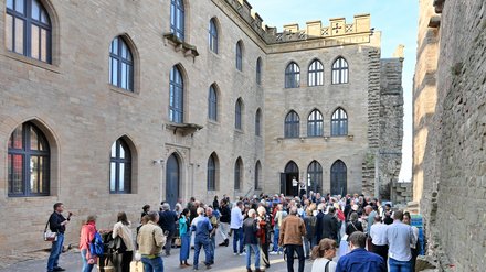 "Große Außenveranstaltung in einem historischen Innenhof mit Backsteingebäuden. Etwa 100 Personen in casual bis Business-Kleidung stehen und gehen durcheinander. Die umgebenden Gebäude zeigen mittelalterliche Architektur mit großen gotischen Fenstern, Zinnen und Steinmauern in Beige und Grau. Sonnenschein bei klarem Himmel beleuchtet die Szene. Links sitzt eine Person mit Tablet und nimmt Notizen."