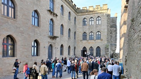 "Große Außenveranstaltung in einem historischen Innenhof mit Backsteingebäuden. Etwa 100 Personen in casual bis Business-Kleidung stehen und gehen durcheinander. Die umgebenden Gebäude zeigen mittelalterliche Architektur mit großen gotischen Fenstern, Zinnen und Steinmauern in Beige und Grau. Sonnenschein bei klarem Himmel beleuchtet die Szene. Links sitzt eine Person mit Tablet und nimmt Notizen."