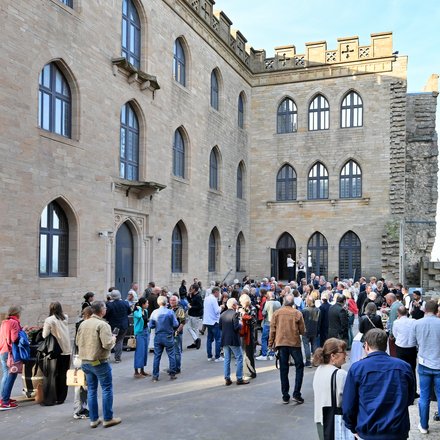 "Große Außenveranstaltung in einem historischen Innenhof mit Backsteingebäuden. Etwa 100 Personen in casual bis Business-Kleidung stehen und gehen durcheinander. Die umgebenden Gebäude zeigen mittelalterliche Architektur mit großen gotischen Fenstern, Zinnen und Steinmauern in Beige und Grau. Sonnenschein bei klarem Himmel beleuchtet die Szene. Links sitzt eine Person mit Tablet und nimmt Notizen."