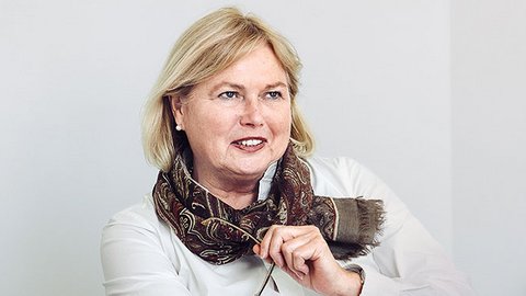 "Porträtfoto einer Frau mittleren Alters mit blondem, schulterlangem Haar und roten Lippen. Sie trägt eine weiße Bluse und einen braun-gemusterten Schal. Sie sitzt an einem Tisch, lehnt sich vor und hält einen Pinsel in der Hand. Lächelnder, selbstbewusster Gesichtsausdruck. Heller grauer Hintergrund. Professionelle Geschäftsfotografie."