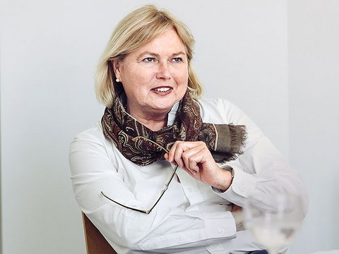 "Porträtfoto einer Frau mittleren Alters mit blondem, schulterlangem Haar und roten Lippen. Sie trägt eine weiße Bluse und einen braun-gemusterten Schal. Sie sitzt an einem Tisch, lehnt sich vor und hält einen Pinsel in der Hand. Lächelnder, selbstbewusster Gesichtsausdruck. Heller grauer Hintergrund. Professionelle Geschäftsfotografie."