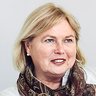 "Porträtfoto einer Frau mittleren Alters mit blondem, schulterlangem Haar und roten Lippen. Sie trägt eine weiße Bluse und einen braun-gemusterten Schal. Sie sitzt an einem Tisch, lehnt sich vor und hält einen Pinsel in der Hand. Lächelnder, selbstbewusster Gesichtsausdruck. Heller grauer Hintergrund. Professionelle Geschäftsfotografie."