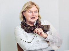 "Porträtfoto einer Frau mittleren Alters mit blondem, schulterlangem Haar und roten Lippen. Sie trägt eine weiße Bluse und einen braun-gemusterten Schal. Sie sitzt an einem Tisch, lehnt sich vor und hält einen Pinsel in der Hand. Lächelnder, selbstbewusster Gesichtsausdruck. Heller grauer Hintergrund. Professionelle Geschäftsfotografie."