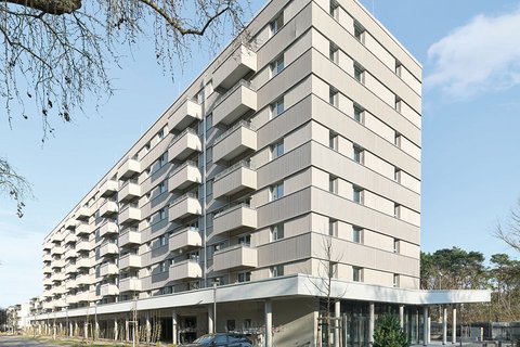 Modernes Mehrfamilienhaus mit neun Geschossen und cremefarbeener Fassade. Das Gebäude hat eine pyramidenförmige Dachform und ist mit regelmäßig angeordneten Balkonen und Fenster an jeder Etage ausgestattet. Im Erdgeschoss befindet sich eine offene Loggia mit großen Glasfenstern und einer Tiefgarage. Ein dunkelblauer PKW parkt vor dem Haus. Der Platz vor dem Gebäude ist gepflastert mit jungen Bäumen und Grünflächen. Links oben ist eine blattlose Trauerweide teilweise sichtbar. Der Himmel ist größtenteils klar und blau mit vereinzelten Wolken.