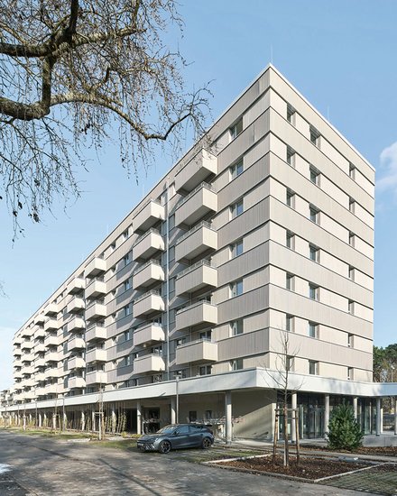 Modernes Mehrfamilienhaus mit neun Geschossen und cremefarbeener Fassade. Das Gebäude hat eine pyramidenförmige Dachform und ist mit regelmäßig angeordneten Balkonen und Fenster an jeder Etage ausgestattet. Im Erdgeschoss befindet sich eine offene Loggia mit großen Glasfenstern und einer Tiefgarage. Ein dunkelblauer PKW parkt vor dem Haus. Der Platz vor dem Gebäude ist gepflastert mit jungen Bäumen und Grünflächen. Links oben ist eine blattlose Trauerweide teilweise sichtbar. Der Himmel ist größtenteils klar und blau mit vereinzelten Wolken.