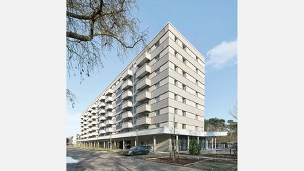 Modernes Mehrfamilienhaus mit neun Geschossen und cremefarbeener Fassade. Das Gebäude hat eine pyramidenförmige Dachform und ist mit regelmäßig angeordneten Balkonen und Fenster an jeder Etage ausgestattet. Im Erdgeschoss befindet sich eine offene Loggia mit großen Glasfenstern und einer Tiefgarage. Ein dunkelblauer PKW parkt vor dem Haus. Der Platz vor dem Gebäude ist gepflastert mit jungen Bäumen und Grünflächen. Links oben ist eine blattlose Trauerweide teilweise sichtbar. Der Himmel ist größtenteils klar und blau mit vereinzelten Wolken.