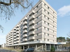 Modernes Mehrfamilienhaus mit neun Geschossen und cremefarbeener Fassade. Das Gebäude hat eine pyramidenförmige Dachform und ist mit regelmäßig angeordneten Balkonen und Fenster an jeder Etage ausgestattet. Im Erdgeschoss befindet sich eine offene Loggia mit großen Glasfenstern und einer Tiefgarage. Ein dunkelblauer PKW parkt vor dem Haus. Der Platz vor dem Gebäude ist gepflastert mit jungen Bäumen und Grünflächen. Links oben ist eine blattlose Trauerweide teilweise sichtbar. Der Himmel ist größtenteils klar und blau mit vereinzelten Wolken.