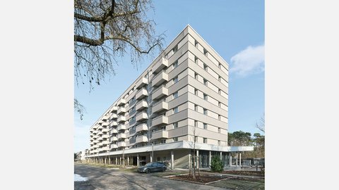 Modernes Mehrfamilienhaus mit neun Geschossen und cremefarbeener Fassade. Das Gebäude hat eine pyramidenförmige Dachform und ist mit regelmäßig angeordneten Balkonen und Fenster an jeder Etage ausgestattet. Im Erdgeschoss befindet sich eine offene Loggia mit großen Glasfenstern und einer Tiefgarage. Ein dunkelblauer PKW parkt vor dem Haus. Der Platz vor dem Gebäude ist gepflastert mit jungen Bäumen und Grünflächen. Links oben ist eine blattlose Trauerweide teilweise sichtbar. Der Himmel ist größtenteils klar und blau mit vereinzelten Wolken.