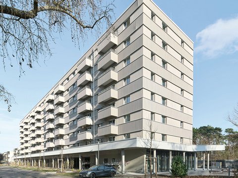 Modernes Mehrfamilienhaus mit neun Geschossen und cremefarbeener Fassade. Das Gebäude hat eine pyramidenförmige Dachform und ist mit regelmäßig angeordneten Balkonen und Fenster an jeder Etage ausgestattet. Im Erdgeschoss befindet sich eine offene Loggia mit großen Glasfenstern und einer Tiefgarage. Ein dunkelblauer PKW parkt vor dem Haus. Der Platz vor dem Gebäude ist gepflastert mit jungen Bäumen und Grünflächen. Links oben ist eine blattlose Trauerweide teilweise sichtbar. Der Himmel ist größtenteils klar und blau mit vereinzelten Wolken.