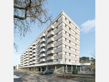 Modernes Mehrfamilienhaus mit neun Geschossen und cremefarbeener Fassade. Das Gebäude hat eine pyramidenförmige Dachform und ist mit regelmäßig angeordneten Balkonen und Fenster an jeder Etage ausgestattet. Im Erdgeschoss befindet sich eine offene Loggia mit großen Glasfenstern und einer Tiefgarage. Ein dunkelblauer PKW parkt vor dem Haus. Der Platz vor dem Gebäude ist gepflastert mit jungen Bäumen und Grünflächen. Links oben ist eine blattlose Trauerweide teilweise sichtbar. Der Himmel ist größtenteils klar und blau mit vereinzelten Wolken.
