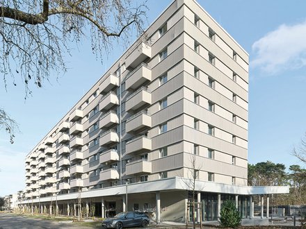 Modernes Mehrfamilienhaus mit neun Geschossen und cremefarbeener Fassade. Das Gebäude hat eine pyramidenförmige Dachform und ist mit regelmäßig angeordneten Balkonen und Fenster an jeder Etage ausgestattet. Im Erdgeschoss befindet sich eine offene Loggia mit großen Glasfenstern und einer Tiefgarage. Ein dunkelblauer PKW parkt vor dem Haus. Der Platz vor dem Gebäude ist gepflastert mit jungen Bäumen und Grünflächen. Links oben ist eine blattlose Trauerweide teilweise sichtbar. Der Himmel ist größtenteils klar und blau mit vereinzelten Wolken.