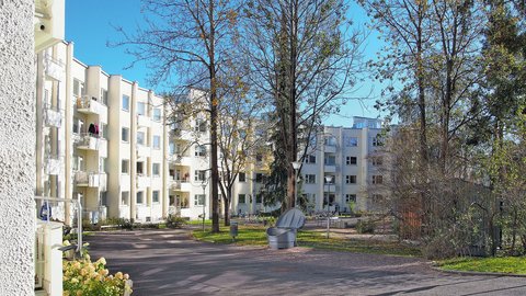 "Blick auf einen ruhigen Innenhofbereich mit mehreren viergeschossigen Wohngebäuden in Creme- und Beigeton. Der gepflasterte Weg wird von Bäumen mit teilweise herbstlichem Laub und Grünanlagen gesäumt. Ein grauer Mülleimer steht in der Bildmitte. Der Himmel ist klar und blau."