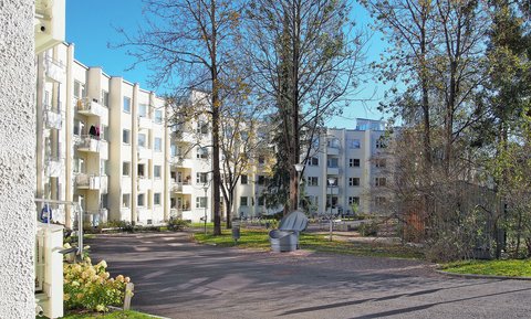 "Blick auf einen ruhigen Innenhofbereich mit mehreren viergeschossigen Wohngebäuden in Creme- und Beigeton. Der gepflasterte Weg wird von Bäumen mit teilweise herbstlichem Laub und Grünanlagen gesäumt. Ein grauer Mülleimer steht in der Bildmitte. Der Himmel ist klar und blau."