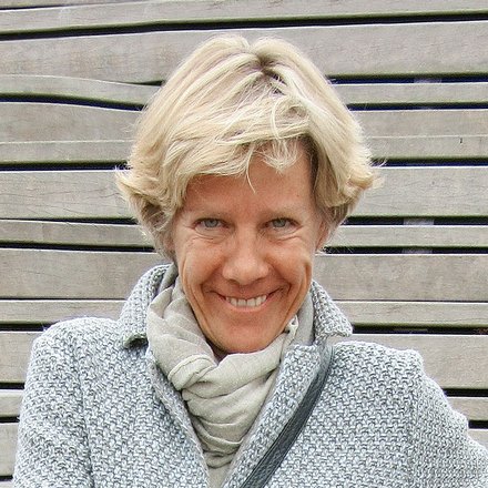 "Porträtfoto einer Frau mit schulterlangem blondem Haar, die auf einer grauen Holzbank sitzt. Sie trägt einen hellgrauen Strickpullover mit Schalkragen und blaue Jeans. Sie lächelt in die Kamera und ruht ihre Arme entspannt auf ihren Beinen. Im Hintergrund ist eine horizontal gestreifte graue Holzwand zu sehen."