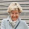 "Porträtfoto einer Frau mit schulterlangem blondem Haar, die auf einer grauen Holzbank sitzt. Sie trägt einen hellgrauen Strickpullover mit Schalkragen und blaue Jeans. Sie lächelt in die Kamera und ruht ihre Arme entspannt auf ihren Beinen. Im Hintergrund ist eine horizontal gestreifte graue Holzwand zu sehen."