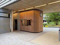 Verkaufsstand aus vertikalen Holzlamellen mit geöffneter Klappe und Uhr an der Außenwand unter einem betonierten Gebäudedurchgang.