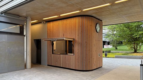 Verkaufsstand aus vertikalen Holzlamellen mit geöffneter Klappe und Uhr an der Außenwand unter einem betonierten Gebäudedurchgang.