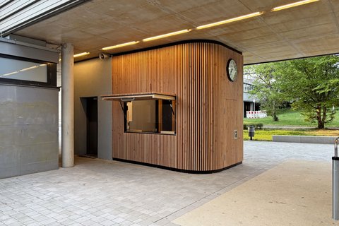 Verkaufsstand aus vertikalen Holzlamellen mit geöffneter Klappe und Uhr an der Außenwand unter einem betonierten Gebäudedurchgang.