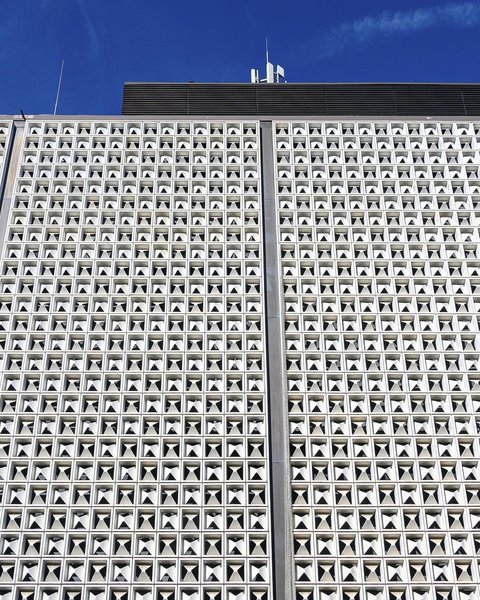 Weiße, gerasterte Fassade eines Flughafengebäudes mit zahlreichen dreieckigen Öffnungen vor blauem Himmel.