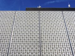 Weiße, gerasterte Fassade eines Flughafengebäudes mit zahlreichen dreieckigen Öffnungen vor blauem Himmel.