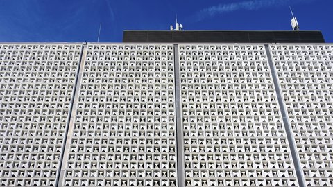 Weiße, gerasterte Fassade eines Flughafengebäudes mit zahlreichen dreieckigen Öffnungen vor blauem Himmel.