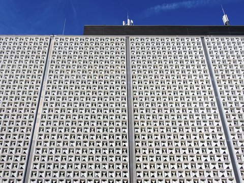 Weiße, gerasterte Fassade eines Flughafengebäudes mit zahlreichen dreieckigen Öffnungen vor blauem Himmel.