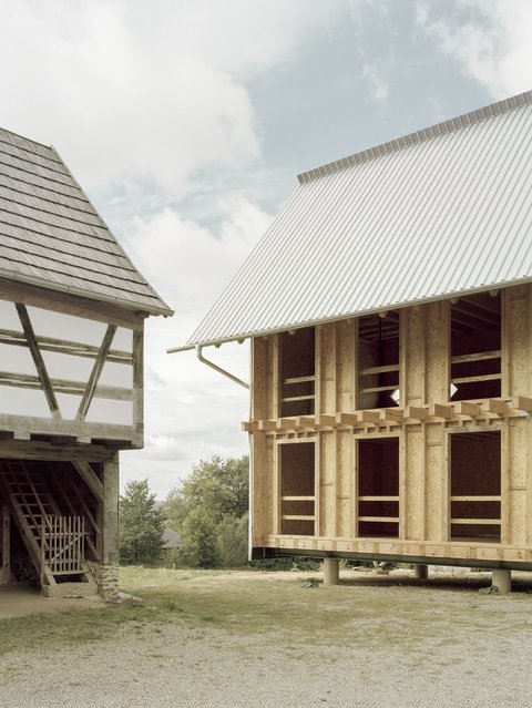 Eine Außenaufnahme im Halbnahbereich, die zwei Gebäude nebeneinander zeigt. Links ist ein historisches Fachwerkgebäude mit dunklen Holzbalken und hellen Gefachen, bedeckt von einem alten Schindeldach. Rechts daneben steht ein modernes, im Bau befindliches Gebäude in Holzrahmenbauweise. Die Wände sind mit OSB-Platten verkleidet und es hat ein großes, helles, gewelltes Metalldach. Das Gebäude ruht auf Betonpfeilern. Der Himmel ist bewölkt.