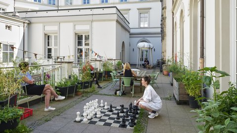 Innenhof mit mehreren Personen, die an Tischen sitzen und eine große Schachfigur auf einem überdimensionalen Schachbrett bewegen
