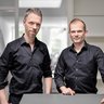 Zwei Männer in schwarzen Hemden stehen nebeneinander in einem hellen Büro und blicken in die Kamera.