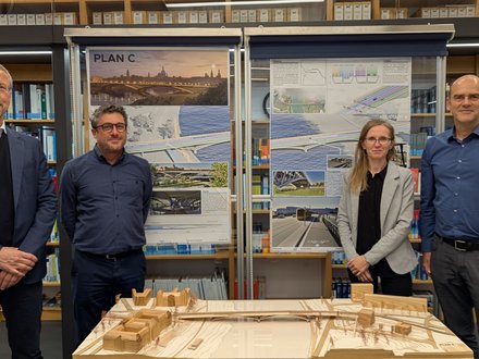 Mehrere Personen stehen in einem Innenraum vor Ausstellungstafeln und einem Holzmodell, das eine Brücke und ihr Umfeld zeigt, die Präsentation ist sachlich und konzentriert, wodurch das Bild die Bewertung und Diskussion eines Entwurfs dokumentiert und für fachlichen Austausch, Wettbewerbsverfahren und kollektive Entscheidungsfindung in der Baukultur steht.