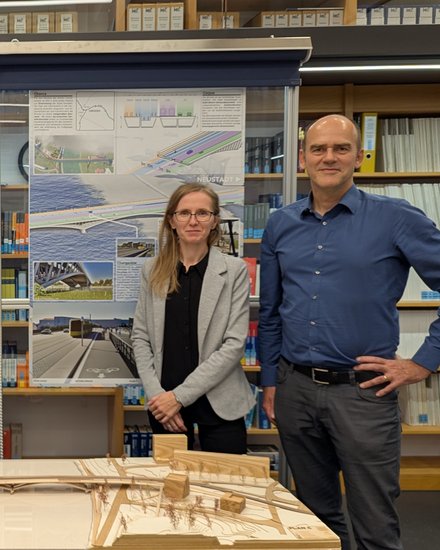 Mehrere Personen stehen in einem Innenraum vor Ausstellungstafeln und einem Holzmodell, das eine Brücke und ihr Umfeld zeigt, die Präsentation ist sachlich und konzentriert, wodurch das Bild die Bewertung und Diskussion eines Entwurfs dokumentiert und für fachlichen Austausch, Wettbewerbsverfahren und kollektive Entscheidungsfindung in der Baukultur steht.