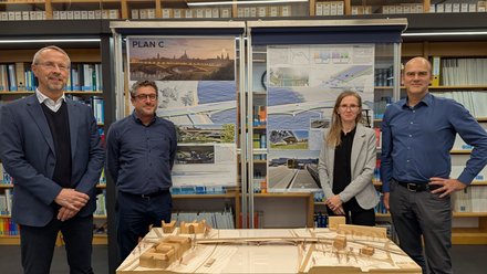 Mehrere Personen stehen in einem Innenraum vor Ausstellungstafeln und einem Holzmodell, das eine Brücke und ihr Umfeld zeigt, die Präsentation ist sachlich und konzentriert, wodurch das Bild die Bewertung und Diskussion eines Entwurfs dokumentiert und für fachlichen Austausch, Wettbewerbsverfahren und kollektive Entscheidungsfindung in der Baukultur steht.