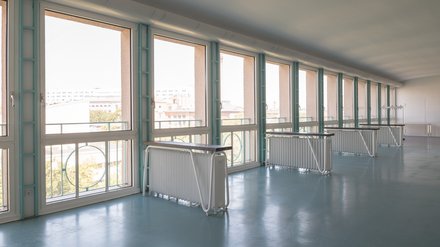 Langer, heller Raum mit durchgehender Fensterfront und türkisfarbenen Rahmen; davor weiße Heizkörper und Pulte, Stadtblick.