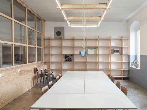 Ein heller, moderner Besprechungs- oder Werkstattraum mit einem langen weißen Tisch im Vordergrund. Der Raum ist geprägt von unbehandeltem Holz: Eine große Holzregalwand bedeckt die Rückseite, und links gibt es eine Trennwand mit vielen Glasscheiben in Holzrahmen. An der Decke hängt ein großer, rechteckiger Holzleuchter mit LED-Röhren und einer herabhängenden Steckdosenampel. Auf der rechten Seite spendet ein bogenförmiges Fenster Tageslicht. Die Einrichtung wirkt minimalistisch und funktional.
