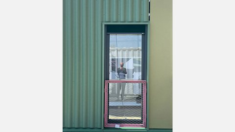 Das Bild zeigt eine Gebäudefassade mit unterschiedlichen Materialien. Links ist eine grünlich gefärbte, gewellte Metallfassade zu sehen, rechts eine glatte, hellere Fläche. In der Mitte befindet sich ein Fenster, in dessen Scheibe sich eine Person spiegelt, die offenbar ein Foto aufnimmt. Vor dem Fenster ist ein temporär montiertes Bauteil mit Metallrahmen und Gitter angebracht. Zweck des Bildes ist es, Details einer Fassade aus wiederverwendeten oder modularen Bauelementen zu zeigen und auf die Materialität sowie die Anpassungsfähigkeit zirkulärer Bauweisen hinzuweisen.