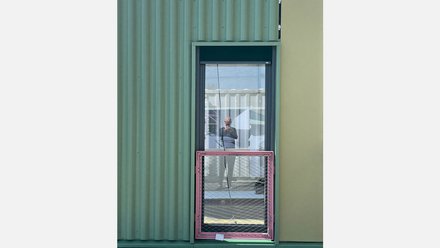 Das Bild zeigt eine Gebäudefassade mit unterschiedlichen Materialien. Links ist eine grünlich gefärbte, gewellte Metallfassade zu sehen, rechts eine glatte, hellere Fläche. In der Mitte befindet sich ein Fenster, in dessen Scheibe sich eine Person spiegelt, die offenbar ein Foto aufnimmt. Vor dem Fenster ist ein temporär montiertes Bauteil mit Metallrahmen und Gitter angebracht. Zweck des Bildes ist es, Details einer Fassade aus wiederverwendeten oder modularen Bauelementen zu zeigen und auf die Materialität sowie die Anpassungsfähigkeit zirkulärer Bauweisen hinzuweisen.