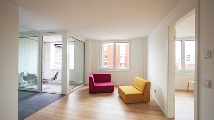 Heller Wohnraum mit Holzfußboden, großen Fenstern und zwei farbigen Sesseln.