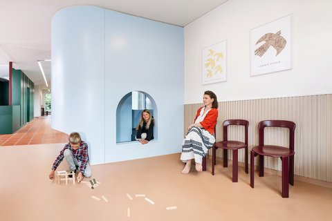 „Ein helles Wartezimmer: Eine Frau sitzt auf einem Holzstuhl, während ein Junge im Vordergrund mit Bauklötzen spielt und ein Mädchen aus der bogenförmigen Öffnung eines hellblauen Spielelements schaut.“