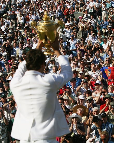  Tennisspieler in weißem Poloshirt hält einen großen goldenen Pokal in die Höhe. Im Hintergrund jubelt eine große Menge von Zuschauern auf den Tribünen. Viele Zuschauer fotografieren und filmen den Moment mit ihren Smartphones. Die Szene spielt sich bei sonnigem Wetter ab.