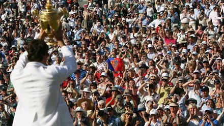  Tennisspieler in weißem Poloshirt hält einen großen goldenen Pokal in die Höhe. Im Hintergrund jubelt eine große Menge von Zuschauern auf den Tribünen. Viele Zuschauer fotografieren und filmen den Moment mit ihren Smartphones. Die Szene spielt sich bei sonnigem Wetter ab.