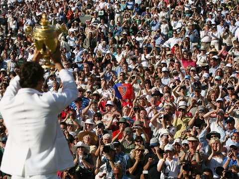  Tennisspieler in weißem Poloshirt hält einen großen goldenen Pokal in die Höhe. Im Hintergrund jubelt eine große Menge von Zuschauern auf den Tribünen. Viele Zuschauer fotografieren und filmen den Moment mit ihren Smartphones. Die Szene spielt sich bei sonnigem Wetter ab.