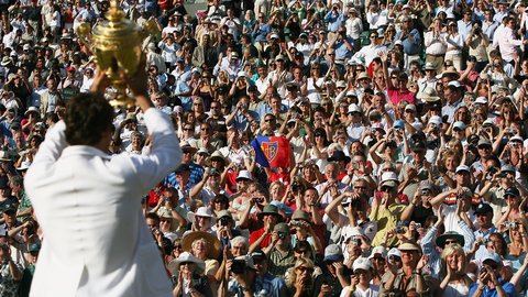  Tennisspieler in weißem Poloshirt hält einen großen goldenen Pokal in die Höhe. Im Hintergrund jubelt eine große Menge von Zuschauern auf den Tribünen. Viele Zuschauer fotografieren und filmen den Moment mit ihren Smartphones. Die Szene spielt sich bei sonnigem Wetter ab.