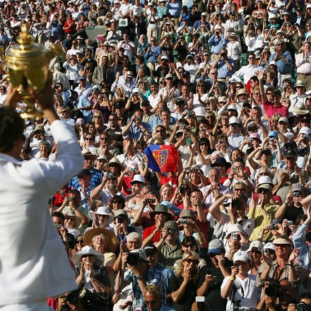  Tennisspieler in weißem Poloshirt hält einen großen goldenen Pokal in die Höhe. Im Hintergrund jubelt eine große Menge von Zuschauern auf den Tribünen. Viele Zuschauer fotografieren und filmen den Moment mit ihren Smartphones. Die Szene spielt sich bei sonnigem Wetter ab.