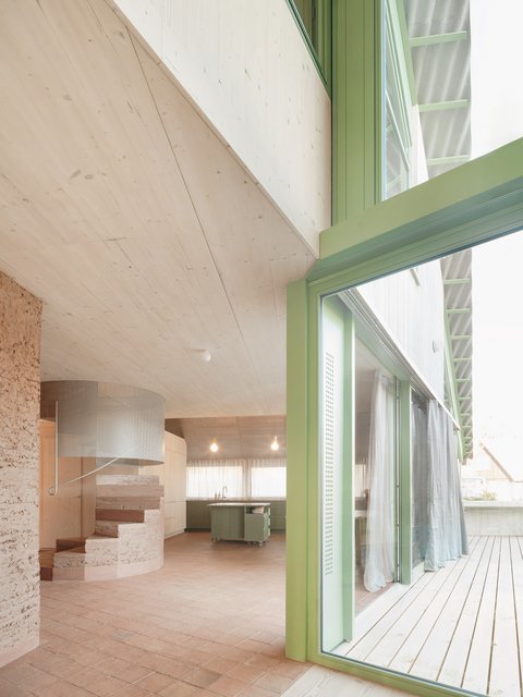 "Großzügiger offener Wohnbereich mit Holzdecke, Holzwänden und grünem Strukturelement. Grüne Küchenzeile mit Holzarbeitsplatte. Heller Terrakotta-Treppen-Element links. Weiße Vorhänge an großen Fenstern."