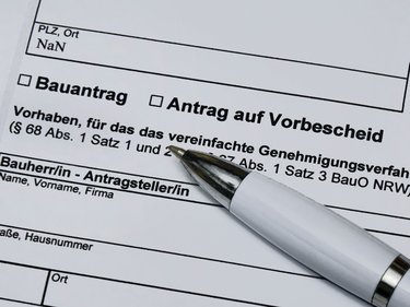 Ein stark vergrößerter Ausschnitt eines offiziellen Formulars zeigt die Felder „Bauantrag“ und „Antrag auf Vorbescheid“ sowie Textpassagen zu einem vereinfachten Genehmigungsverfahren nach der Bauordnung, ein weißer Kugelschreiber liegt schräg auf dem Papier und weist auf den auszufüllenden Bereich, wodurch das Bild administrative Abläufe rund um Planen und Bauen sichtbar macht und für die formalen, rechtlichen Rahmenbedingungen steht, die Baukultur und bauliche Umsetzung begleiten.