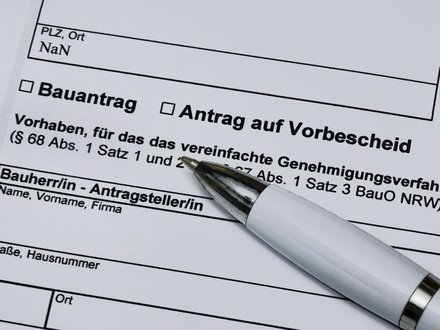 Ein stark vergrößerter Ausschnitt eines offiziellen Formulars zeigt die Felder „Bauantrag“ und „Antrag auf Vorbescheid“ sowie Textpassagen zu einem vereinfachten Genehmigungsverfahren nach der Bauordnung, ein weißer Kugelschreiber liegt schräg auf dem Papier und weist auf den auszufüllenden Bereich, wodurch das Bild administrative Abläufe rund um Planen und Bauen sichtbar macht und für die formalen, rechtlichen Rahmenbedingungen steht, die Baukultur und bauliche Umsetzung begleiten.