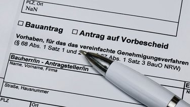 Ein stark vergrößerter Ausschnitt eines offiziellen Formulars zeigt die Felder „Bauantrag“ und „Antrag auf Vorbescheid“ sowie Textpassagen zu einem vereinfachten Genehmigungsverfahren nach der Bauordnung, ein weißer Kugelschreiber liegt schräg auf dem Papier und weist auf den auszufüllenden Bereich, wodurch das Bild administrative Abläufe rund um Planen und Bauen sichtbar macht und für die formalen, rechtlichen Rahmenbedingungen steht, die Baukultur und bauliche Umsetzung begleiten.